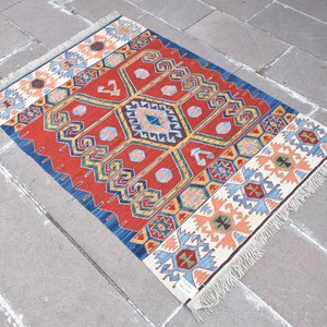 May include: A rectangular, hand-woven rug with a vibrant geometric design. The rug features a red central field with a large diamond pattern in shades of blue, yellow, and peach. The borders are blue and cream with repeating geometric motifs. The rug has fringed edges.