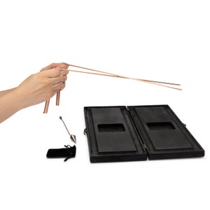 Puede incluir: Un juego de varillas de zahor&iacute; de cobre con dos varillas en forma de L, un p&eacute;ndulo y una caja de madera. La caja est&aacute; abierta, revelando un interior oscuro. Tambi&eacute;n se incluye una peque&ntilde;a bolsa de terciopelo negro.