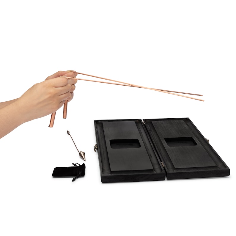 Puede incluir: Un juego de varillas de zahor&iacute; de cobre con dos varillas en forma de L, un p&eacute;ndulo y una caja de madera. La caja est&aacute; abierta, revelando un interior oscuro. Tambi&eacute;n se incluye una peque&ntilde;a bolsa de terciopelo negro.