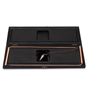 Puede incluir: Caja de madera negra abierta con marco cobrizo y p&eacute;ndulo plateado. El p&eacute;ndulo cuelga de una cadena y se guarda en un compartimento forrado de terciopelo. La caja es rectangular con tapa abisagrada.