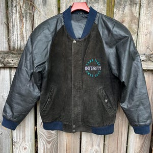 Peut inclure: Veste en cuir et daim noirs avec col, poignets et taille bleu marine. La veste porte l'inscription "CODE ZERO INTENSITY" en lettres turquoise sur la poitrine. La veste a une fermeture à boutons et deux poches avant.