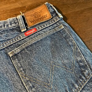 May include: A pair of blue denim jeans with a back pocket featuring a yellow stitching design. The jeans have a brown leather patch with the text "Premium Quality Authentic Wrangler Made in the USA".