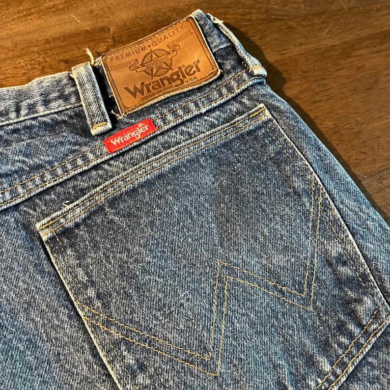 May include: A pair of blue denim jeans with a back pocket featuring a yellow stitching design. The jeans have a brown leather patch with the text "Premium Quality Authentic Wrangler Made in the USA".