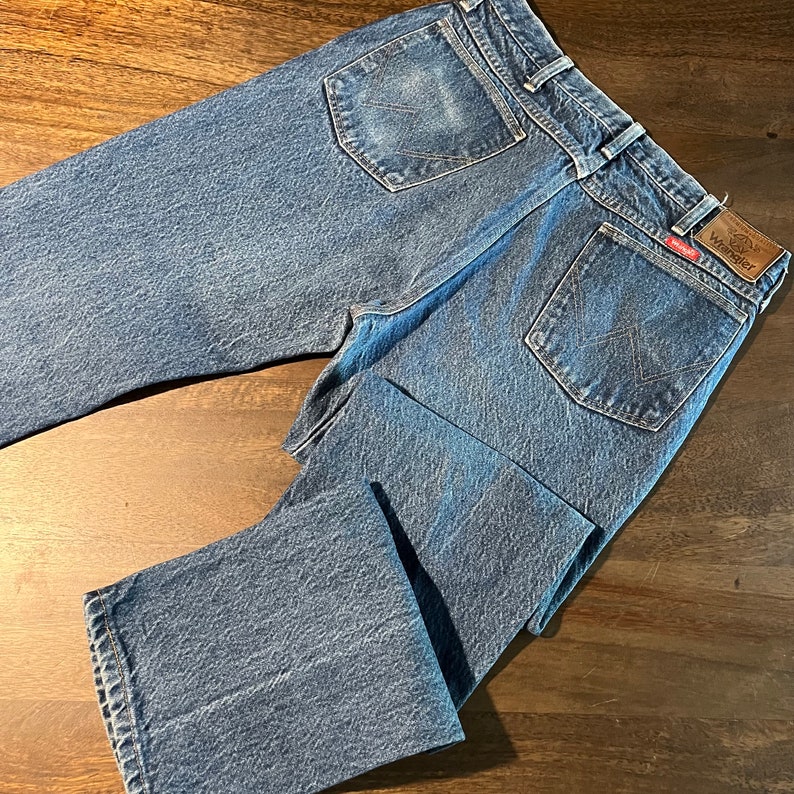 May include: A pair of blue denim jeans with a brown leather Wrangler label on the back pocket.