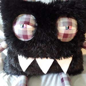 Puede incluir: Un juguete de peluche de monstruo con un cuerpo negro y peludo, ojos grandes a cuadros y dientes triangulares blancos. Los cuernos y las extremidades son de tela a cuadros a juego. El juguete está diseñado para ser suave y tierno.