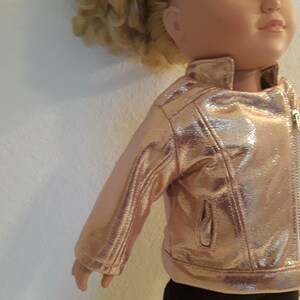 May include: A doll wearing a rose gold faux leather jacket with a zip closure. The jacket has a shiny finish and a pocket on the left side.