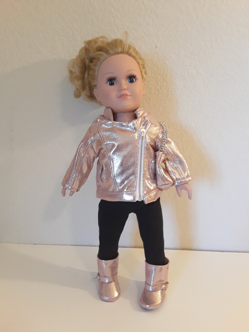 May include: A doll wearing a rose gold jacket, black leggings, and rose gold boots. The doll has blonde hair and blue eyes.