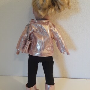 May include: A doll wearing a pink metallic jacket, black leggings, and pink metallic boots. The doll has blonde hair and is carrying a silver chain purse.