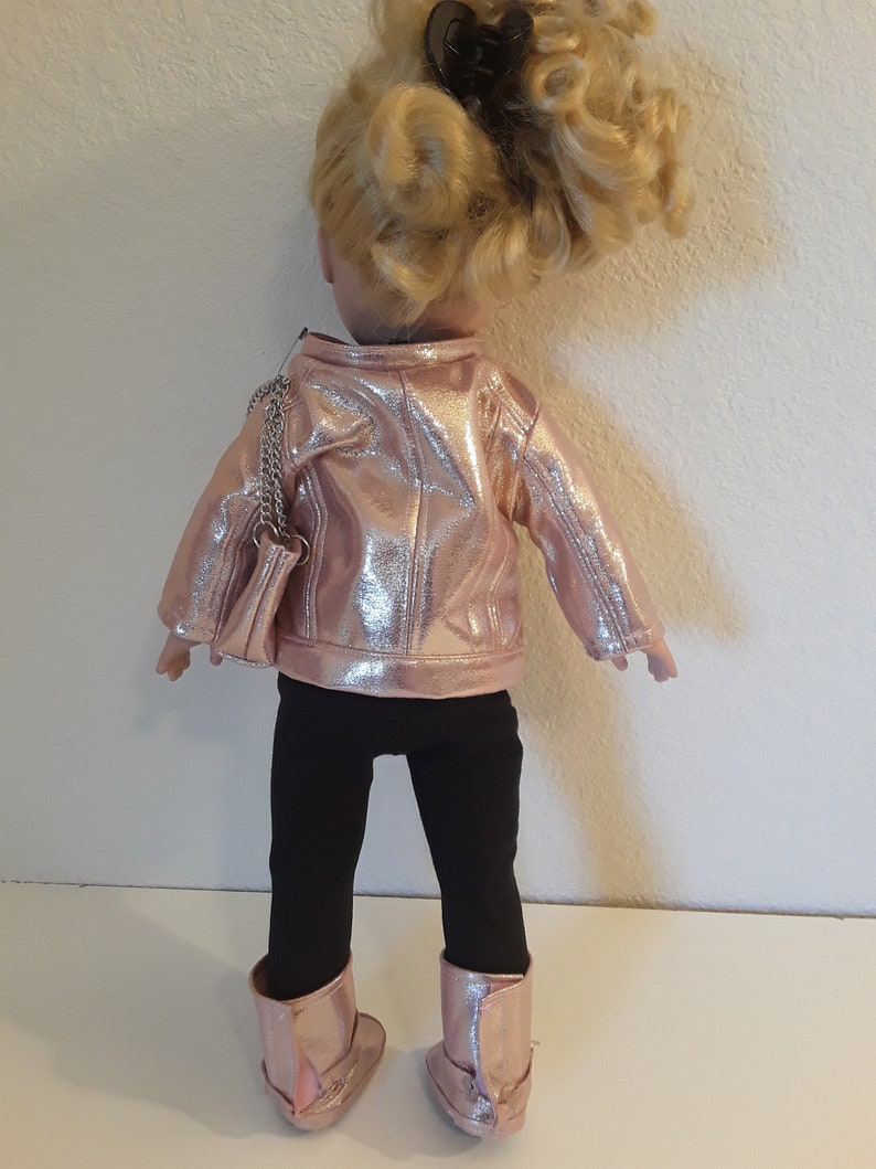 May include: A doll wearing a pink metallic jacket, black leggings, and pink metallic boots. The doll has blonde hair and is carrying a silver chain purse.