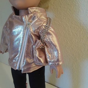 May include: A doll wearing a rose gold jacket, black leggings, and rose gold boots. The doll is holding a rose gold purse with a silver chain strap.