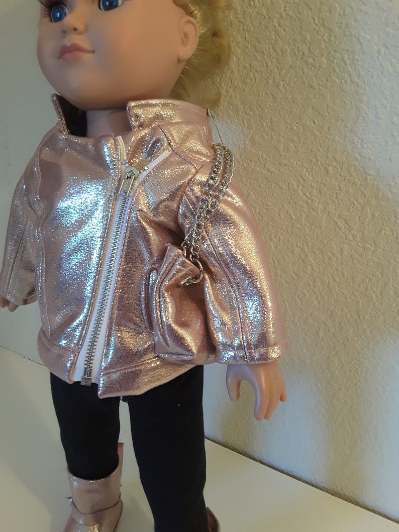 May include: A doll wearing a rose gold jacket, black leggings, and rose gold boots. The doll is holding a rose gold purse with a silver chain strap.