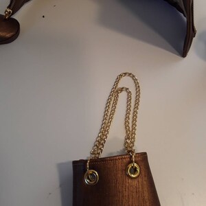 May include: A small brown purse with a gold chain strap. The purse has a textured fabric and two gold rings on the top.