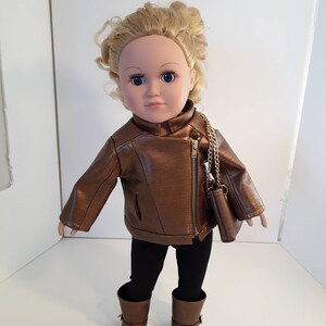 May include: A doll wearing a brown leather jacket, black leggings, and brown boots. The doll has blonde curly hair and blue eyes. The doll is holding a small brown purse with a gold chain.