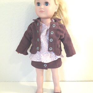 Puede incluir: Una muñeca con una chaqueta marrón, una blusa rosa con estampado floral, una falda marrón y zapatos marrones.