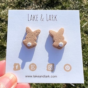 May include: A pair of brown bunny-shaped earrings with white noses. The earrings are displayed on a white card with the text "LAKE & LARK" and a website address.