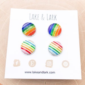 May include: Four round stud earrings with rainbow striped designs on a white card. The card has the text "LAKE & LARK" and social media icons. The website address www.lakeandlark.com is also visible.