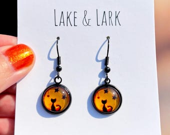 May include: A pair of black dangle earrings with a round, orange glass charm featuring a black silhouette of a cat sitting in front of a star-filled sky. The earrings are labeled "LAKE & LARK".