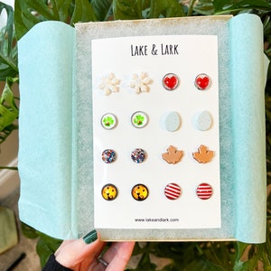 May include: A white card with the text "LAKE & LARK" and the website "www.lakeandlark.com" at the bottom. The card displays a variety of colorful stud earrings with different designs, including snowflakes, hearts, shamrocks, eggs, maple leaves, cats, and stripes.