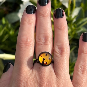 May include: A black ring with a yellow and orange glass cabochon featuring a black silhouette of a cat sitting in front of a crescent moon with stars.