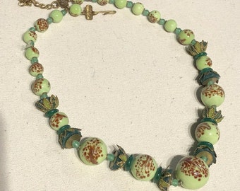 May include: A vintage necklace featuring light green, speckled beads with brown accents. The necklace includes smaller green beads and decorative metal elements. The clasp and chain are gold-toned, creating a unique and elegant accessory.
