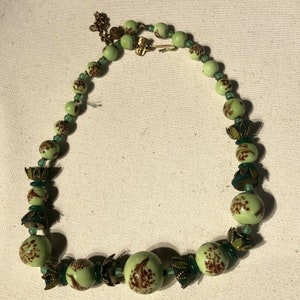 K&ouml;nnte beinhalten: Eine Vintage-Halskette mit hellgr&uuml;nen Perlen mit braunen Blumenmustern. Die Halskette enth&auml;lt kleinere gr&uuml;ne Perlen und dekorative Metallelemente. Der Verschluss ist goldfarben. Ein einzigartiges Schmuckst&uuml;ck.