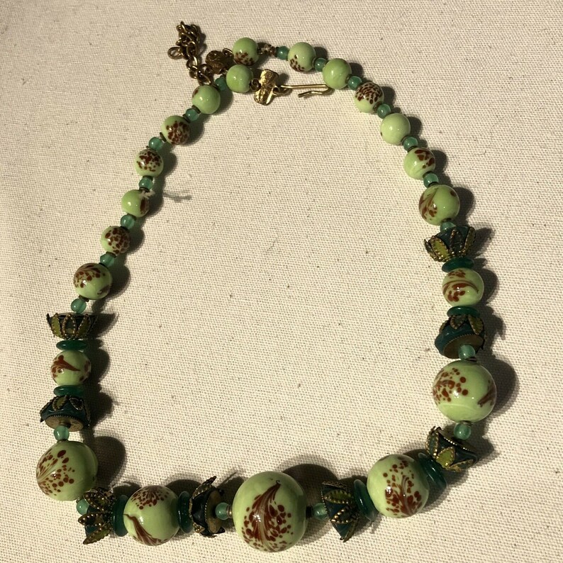 K&ouml;nnte beinhalten: Eine Vintage-Halskette mit hellgr&uuml;nen Perlen mit braunen Blumenmustern. Die Halskette enth&auml;lt kleinere gr&uuml;ne Perlen und dekorative Metallelemente. Der Verschluss ist goldfarben. Ein einzigartiges Schmuckst&uuml;ck.