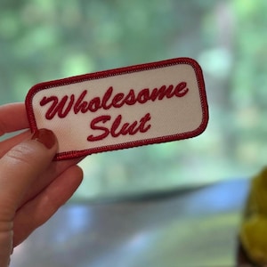 May include: A rectangular patch with a white background and a red border. The words "Wholesome Slut" are embroidered in red cursive script. The patch is held up by a hand with red nail polish, with other patches visible in the background.