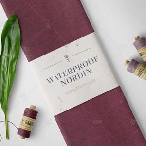 Op de afbeelding: Een bordeauxrode stoffen rol met een wit label met de tekst "WATERPROOF NORDIN 100% POLYESTER". Een groen blad en drie spoelen bordeauxrood garen met bruine labels zijn ook aanwezig. Naai- of knutselmaterialen.