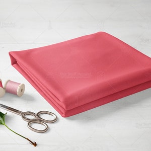 Pode incluir: Um pedaço de tecido rosa dobrado sobre uma superfície de madeira branca. Há dois carretéis de fio e um par de tesouras prateadas ao lado do tecido.