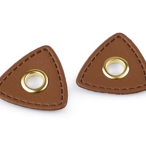 Könnte beinhalten: Zwei braune, dreieckige Lederflicken mit goldenen Ösen. Die Flicken haben einen genähten Rand.
