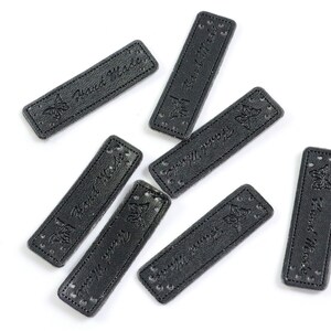 Könnte beinhalten: Sieben schwarze Leder-Etiketten mit dem Text "Hand Made" und einem Schmetterlingsdesign. Die Etiketten sind an den Rändern vernäht und haben Löcher zum Anbringen.