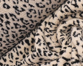 May include: A plush, cream-colored fabric featuring a repeating black leopard print pattern. The material has a thick, textured, fleece-like surface with a soft, fuzzy appearance, shown folded and draped to highlight its dense, tactile pile.