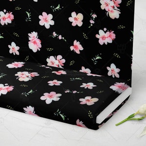 Op de afbeelding: Zwarte stof met een herhalend patroon van roze en witte aquarel kersenbloesems en groene bladeren. De stof is gevouwen en toont het patroon op de boven- en zijkanten. De achtergrond is wit.