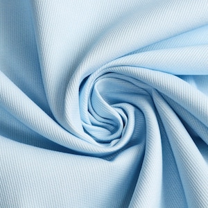 Op de afbeelding: Close-up van een lichtblauwe stof met een subtiele ribtextuur. De stof is in een spiraalvorm gerangschikt, waardoor een wervelend patroon ontstaat. De zachte pastelkleur en de manier waarop de stof is gedrapeerd, suggereren een delicaat materiaal.