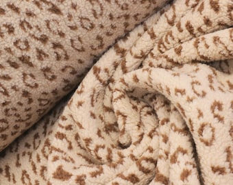May include: A thick, textured fabric featuring a cream-colored base with a repeating brown leopard print pattern. The material has a soft, fuzzy, fleece-like surface with visible folds and drapes that create depth and shadows across the textile.