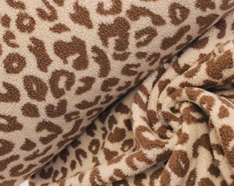 May include: A soft, textured fabric featuring a repeating brown leopard print pattern on a cream-colored background. The material has a dense, fuzzy pile surface with visible folds and drapes, showcasing the animal-inspired design throughout.
