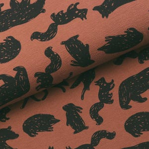 Op de afbeelding: Roestkleurige stof met een herhalend patroon van zwarte dierensilhouetten, waaronder beren, eekhoorns en egels. De stof is gevouwen, wat textuur en diepte laat zien. Ideaal voor knutselprojecten of kleding.