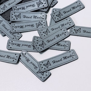 Könnte beinhalten: Ein Stapel grauer Leder-Etiketten mit einem Schmetterlingsdesign und dem aufgedruckten Text "Hand Made".