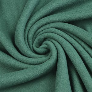 Op de afbeelding: Close-up van een werveling van zachte, groene fleece stof. De stof heeft een getextureerd oppervlak en is strak opgerold in het midden, met plooien die naar buiten uitstralen. De kleur is een gedempt, aards groen.