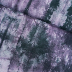 Puede incluir: Primer plano de una tela con un patrón tie-dye en tonos morado, blanco y verde azulado oscuro. La tela está doblada, mostrando la textura y las variaciones de color del tinte. El patrón tiene una apariencia vertical y rayada.