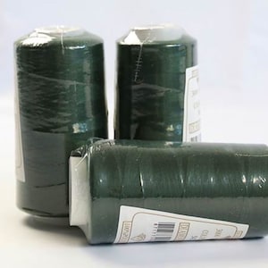 Könnte beinhalten: Drei Spulen mit dunkelgrünem Garn. Die Spulen sind in durchsichtige Plastikfolie verpackt und haben Etiketten mit Text in einer Fremdsprache.