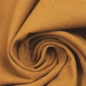 Op de afbeelding: Close-up van een goudbruine canvas stof met een getextureerde weefsel. De stof is gedrapeerd en gewerveld, waardoor zachte plooien en schaduwen ontstaan. De kleur is een warme, aardse tint, geschikt voor diverse knutsel- en designprojecten.