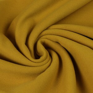 Op de afbeelding: Een close-up van een zachte, mosterdgele stof. De stof is gevouwen en gedrapeerd, waardoor de textuur en de drape zichtbaar worden.