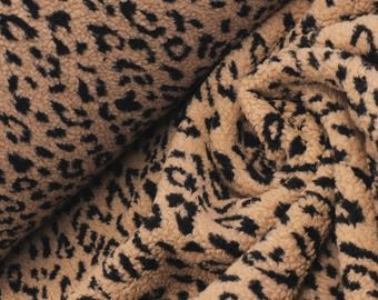 May include: A plush, textured fabric featuring a tan base color with a repeating black leopard print pattern. The material has a soft, fuzzy surface with visible pile, shown in a folded arrangement that creates deep shadows and highlights.