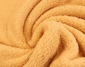 May include: A close-up view of a soft, golden-yellow fabric with a thick, textured sherpa fleece pile. The material is gathered into deep, swirling folds, highlighting the dense, curly, and fuzzy surface characteristic of this synthetic textile.