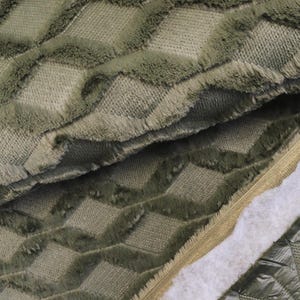 May include: Close-up of a textured fabric in shades of green. The fabric features a repeating diamond pattern with a fuzzy, raised texture. The edges of the diamonds have a fringed appearance. The fabric is folded, revealing layers and a contrasting quilted material.