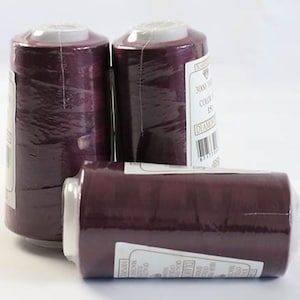 May include: Three spools of dark purple thread, wrapped in clear plastic. Each spool has a white label with text and a barcode. The spools are cylindrical with a white top and bottom.