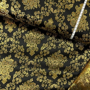 Può includere: Un tessuto nero caratterizzato da un motivo floreale broccato in oro metallizzato. Un metro a nastro bianco è visibile in alto a destra, con segni in centimetri per indicare la scala dell'intricato disegno tessuto sul materiale.