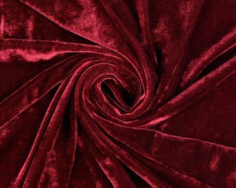 May include: A deep burgundy velvet fabric featuring a dense, soft pile with a light-reflecting sheen. The material is arranged in a central spiral swirl, creating natural folds and shadows that highlight the rich, dark red color and texture.