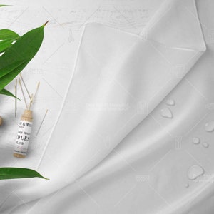 Op de afbeelding: Een witte stof met waterdruppels erop. Een klein houten flesje met een etiket waarop "Knit & Mill" en "Hand Twisted Needles" staat, ligt op een wit oppervlak met groene bladeren.